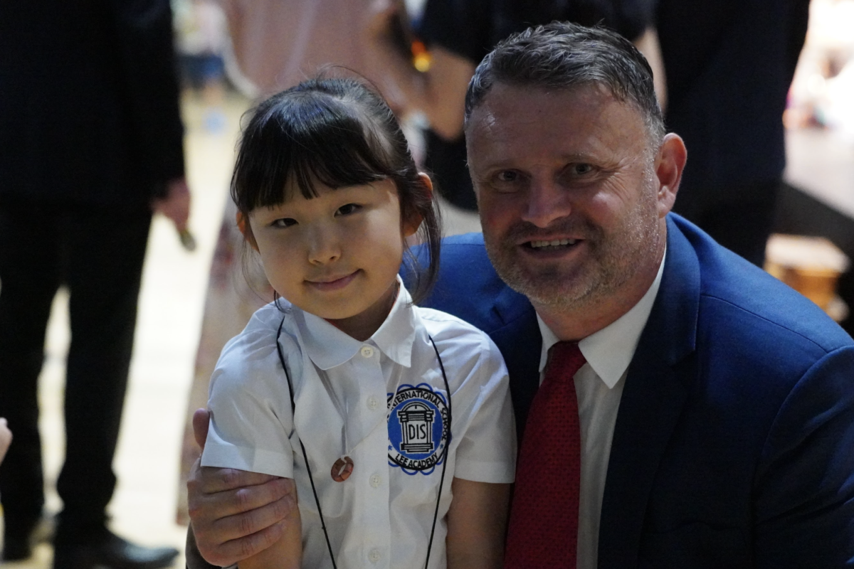Head of school Mr. Jolly congratulates Arin Son in first grade for her hard work. She puts in her handmade necklace to proudly showcase her efforts. 